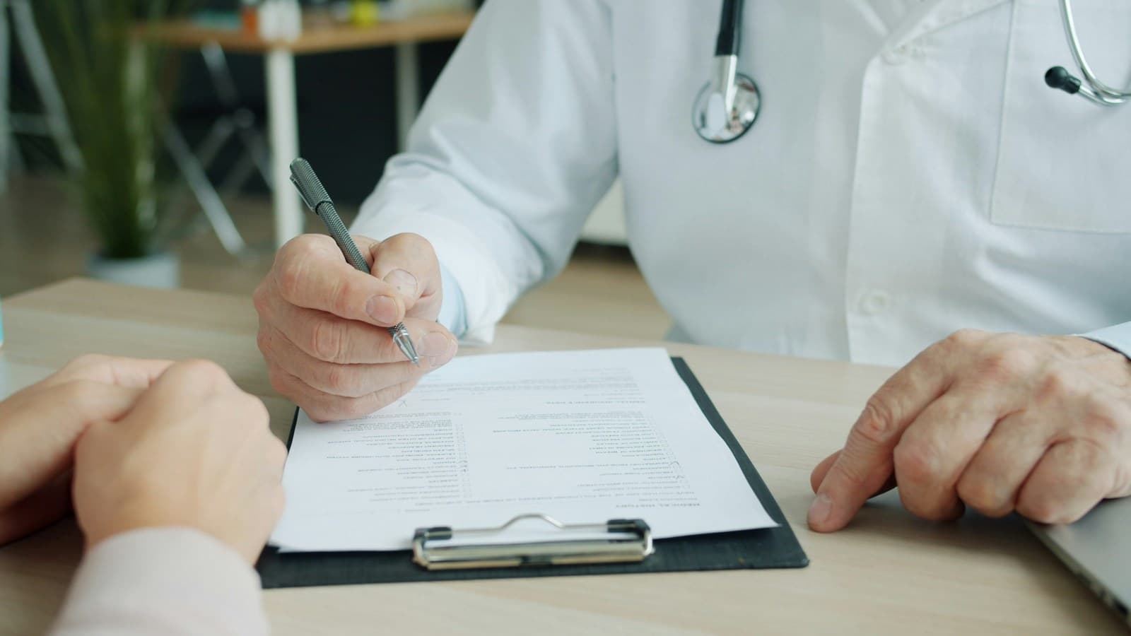 A doctor fills out a patient's medical history form on a clipboard, across the desk from the patient.